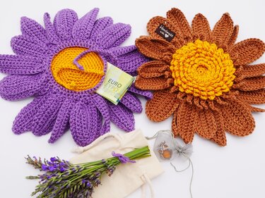 Zwei gehäkelte Blumen (lila und braun), gefalteter Geldschein, Lavendel und Schmuckbeutel auf weißem Grund.