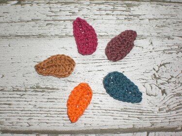 Fünf gehäkelte Blätter in Magenta, Braunrot, Orange, Petrol und Karamell auf weiß getünchtem Holz.