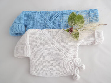 Zwei handgestrickte Wickeljacken für Babys in Hellblau und Weiß, weiße Jacke mit Bindebändern und Bommeln
