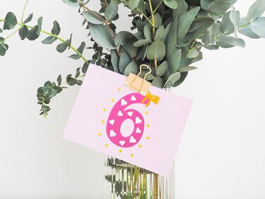 Rosa Karte mit pinker Zahl 6, verziert mit weißen Herzen und gelben Sternen, befestigt mit goldfarbenem Clip an einer Vase mit Eukalyptus