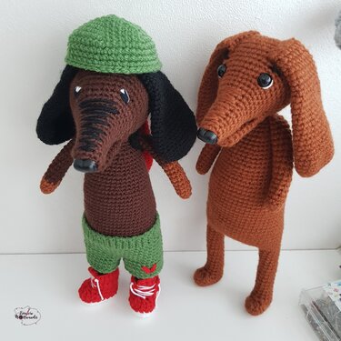 Zwei gehäkelte Dackel-Amigurumi in Braun, links mit grüner Mütze und roten Schuhen, rechts schlicht braun