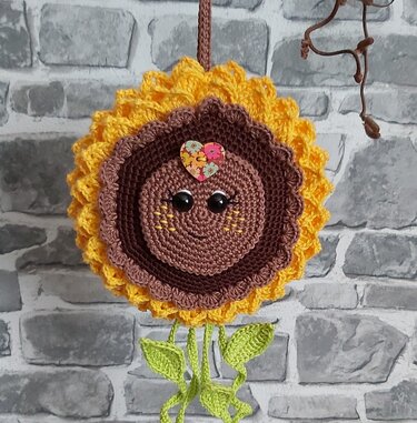 Gehäkelte Sonnenblume in Gelb und Braun mit lächelndem Gesicht, herzförmiger Applikation und hängenden grünen Blättern an grauer Ziegelwand.