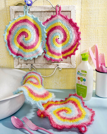 Vier gehäkelte Spiralelemente (Topflappen und Spültücher) in Pastellfarben, zwei hängend, zwei auf einer blauen Arbeitsfläche.