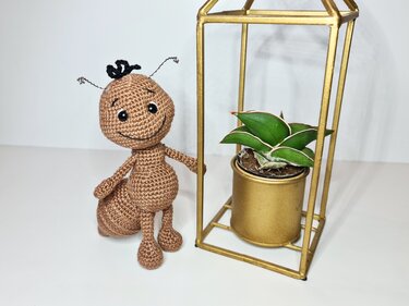 Gehäkelte braune Amigurumi-Ameise mit lächelndem Gesicht und Drahtfühlern neben goldener Pflanzen-Deko