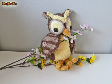 Gehäkelte Eule in Braun, Beige und Gelb mit schwarzen Augen und orangefarbenem Schnabel, umgeben von künstlichen Blüten