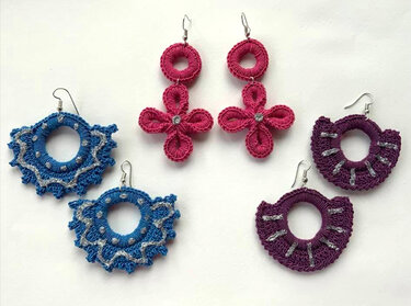 Drei Paar kunstvoll gehäkelte Ohrringe in Pink, Blau und Lila mit metallischen und glitzernden Akzenten, auf einer flachen Oberfläche präsentiert.