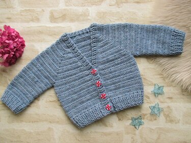 Hellblaue gehäkelte Babyjacke mit langen Ärmeln und vier gemusterten Knöpfen, liegend auf hellem Untergrund mit Deko.