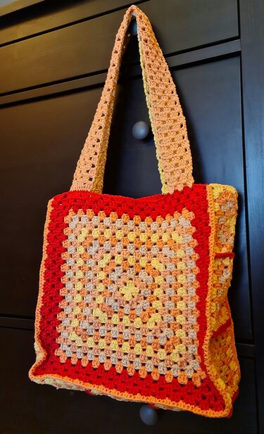 Quadratische gehäkelte Umhängetasche mit Granny‑Square‑Muster in Gelb, Orange und Rot, hängend an dunklem Schrank