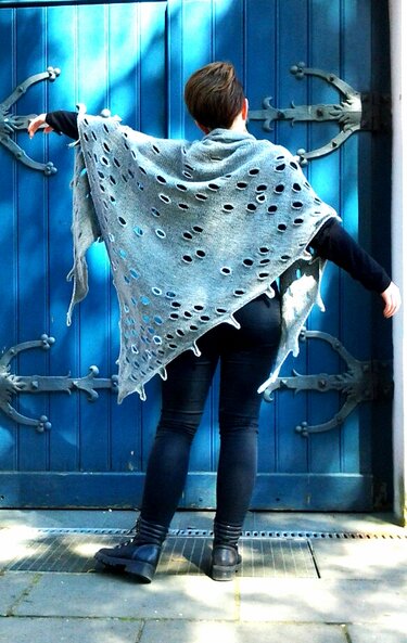 Person von hinten trägt ein graues, grob gestricktes Dreieckstuch mit vielen Lochöffnungen vor einer blauen Tür.