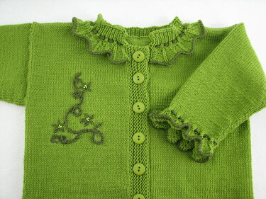 Nahaufnahme einer grünen gestrickten Babyjacke mit Rüschenkragen, Knöpfen und gesticktem Rankenmotiv.