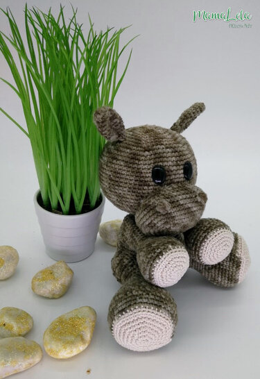 Gehäkeltes Nilpferd-Amigurumi in Braun-Grau mit schwarzen Augen neben einem Topf mit Gras und Dekosteinen auf hellem Hintergrund.