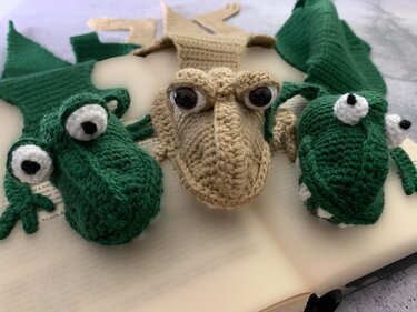 Three crocheted crocodile head bookmarks, two green and one beige, with eye details on an open book
