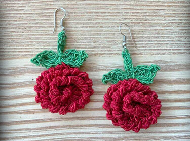 Zwei gehäkelte Ohrringe in Form roter Blüten mit grünen Blättern und silberfarbenen Ohrhaken auf hellem Holz.