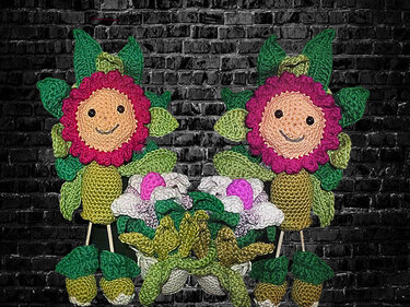 Zwei bunte, handgehäkelte Blumen mit lächelnden Gesichtern, vielen grünen Blättern und kleinen Häkelblüten vor dunkler Ziegelwand