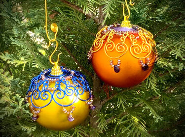Zwei handverzierte Weihnachtskugeln (gelb-blau und orange-gold) mit Perlen an einem Zweig hängend.