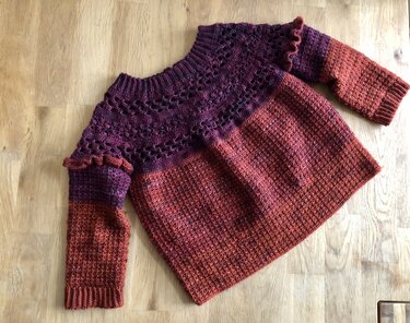 Gehäkelter Kinderpullover in Dunkelviolett und Rostrot mit Lochmuster am Oberteil und Rüschen an den Ärmeln, liegend auf einem Holztisch.