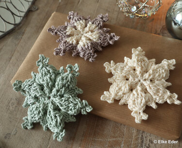 Three crocheted snowflake ornaments in cream, mint green, and two-tone beige on kraft-wrapped paper and a wooden table.