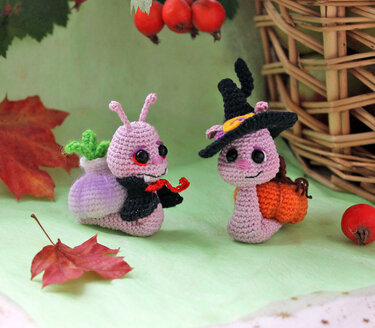 Two crocheted Halloween snail amigurumi: one in a vampire cloak, the other in a witch hat with a pumpkin.