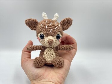 Hand holding small brown and cream crocheted amigurumi deer with black eyes