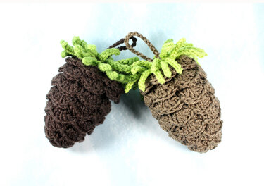 Two crocheted pinecone ornaments, one dark brown and one tan, with green curled leaf tops and a braided hanging loop on a light background.
