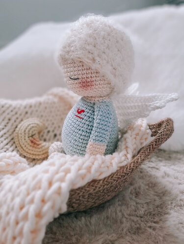 Gehäkeltes Amigurumi-Engelchen mit weißen Flügeln und rotem Herz im Korb auf cremefarbener Decke, mit kleinem Mond