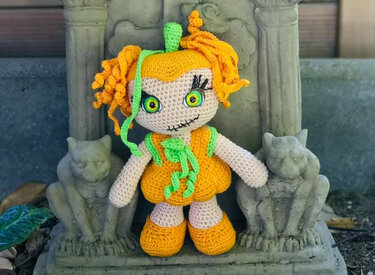 Gehäkelte Amigurumi-Puppe im orange-grünen Kürbis-Look mit großen Augen und gelockten Haaren zwischen zwei steinernen Katzenstatuen