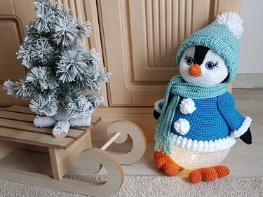 Beleuchteter gehäkelter Pinguin in blauer Jacke und mintgrüner Mütze neben verschneitem Mini-Weihnachtsbaum auf Holzschlitten