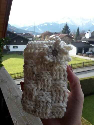Nahaufnahme eines cremefarbenen gehäkelten Utensilos mit Zierblume, vor Häusern und Bergen gehalten.