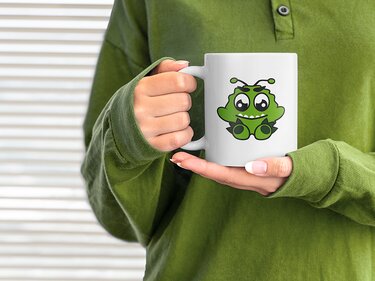 Weiße Tasse mit grünem Cartoon-Außerirdischen, gehalten von Händen in grünem Pullover