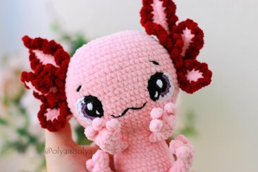 Close-up of a pink crocheted axolotl amigurumi with red gill frills, held by a hand.