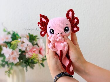 Hand hält rosa gehäkeltes Axolotl-Amigurumi mit roten Kiemen vor hellem Hintergrund