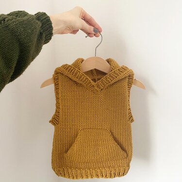 Gestrickte senfgelbe Kinderweste mit Kapuze und Kängurutasche an Holzkleiderbügel
