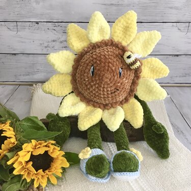 Gehäkelte Sonnenblume-Amigurumi mit gelben Blütenblättern, braunem Gesicht, grünen Beinen, blauen Schuhen und kleiner Biene