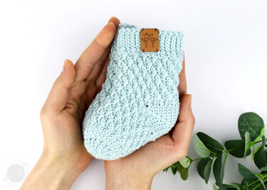 Hands holding a light blue crocheted baby sock with a textured stitch pattern and a small brown bird tag on the ribbed cuff.