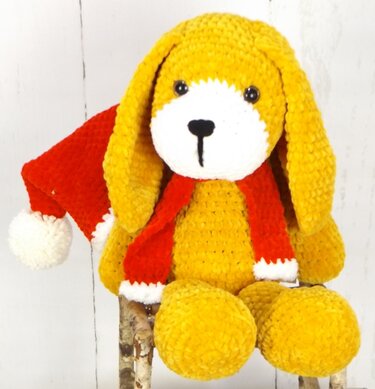 Gehäkelter gelber Amigurumi-Hund mit langen Schlappohren, weißer Schnauze, schwarzen Augen und Nase, roter Weihnachtsmütze mit Pompon und rotem Schal vor hellem Hintergrund.