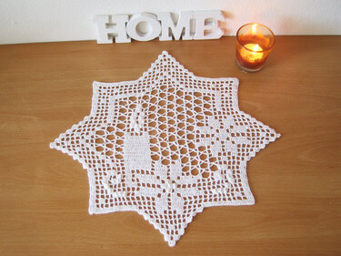 Weißes gehäkeltes Stern-Deckchen mit Weihnachtsmotiven, neben einer brennenden Kerze und einem HOME‑Schriftzug auf einem Holztisch.