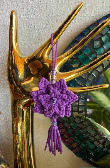 Gehäkelter lila Blumenanhänger mit Perlen und Quaste an einer goldenen Deko-Hand