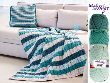 Gestrickte Decke und quadratisches Kissen in Türkis-, Blau- und Weißstreifen auf hellem Sofa; drei Garnknäuel daneben.
