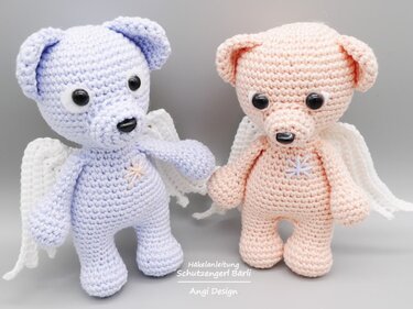 Zwei gehäkelte Teddybären mit weißen Flügeln, einer hellblau, einer hellrosa, mit gestickten Sternen auf der Brust vor grauem Hintergrund.