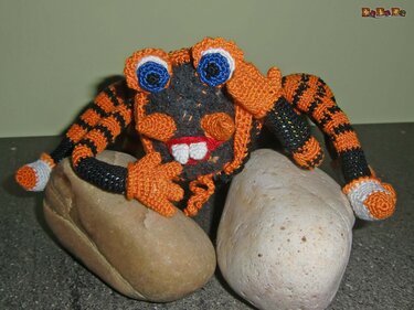 Gehäkelte orange‑schwarze Amigurumi‑Kreatur mit blauen Augen auf Steinen