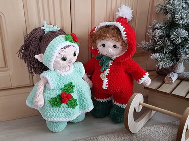 Zwei gehäkelte Amigurumi-Puppen in Weihnachtskleidung, eine mintgrün mit Stechpalmen-Motiv, eine rot mit Kapuze und weißem Rand, daneben ein kleiner Baum