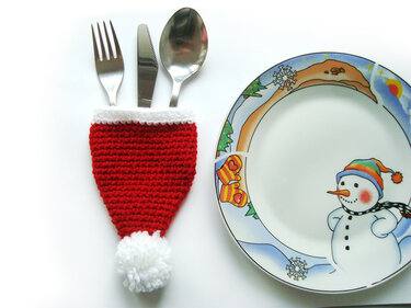 Roter gehäkelter Besteckhalter in Form einer Weihnachtsmütze mit Gabel, Messer und Löffel, daneben ein Teller mit Schneemannmotiv.