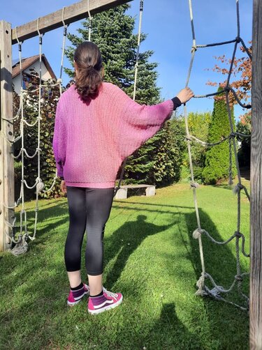 Person von hinten mit pink-lila Ombré-Pullover und weiten Ärmeln, steht auf einer grünen Wiese und hält ein Seilnetz.