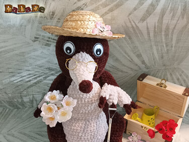 Amigurumi-Maulwurf in Braun und Creme mit Strohhut und Brille, dekoriert mit Blumenstrauß, Spaten und kleiner Gießkanne
