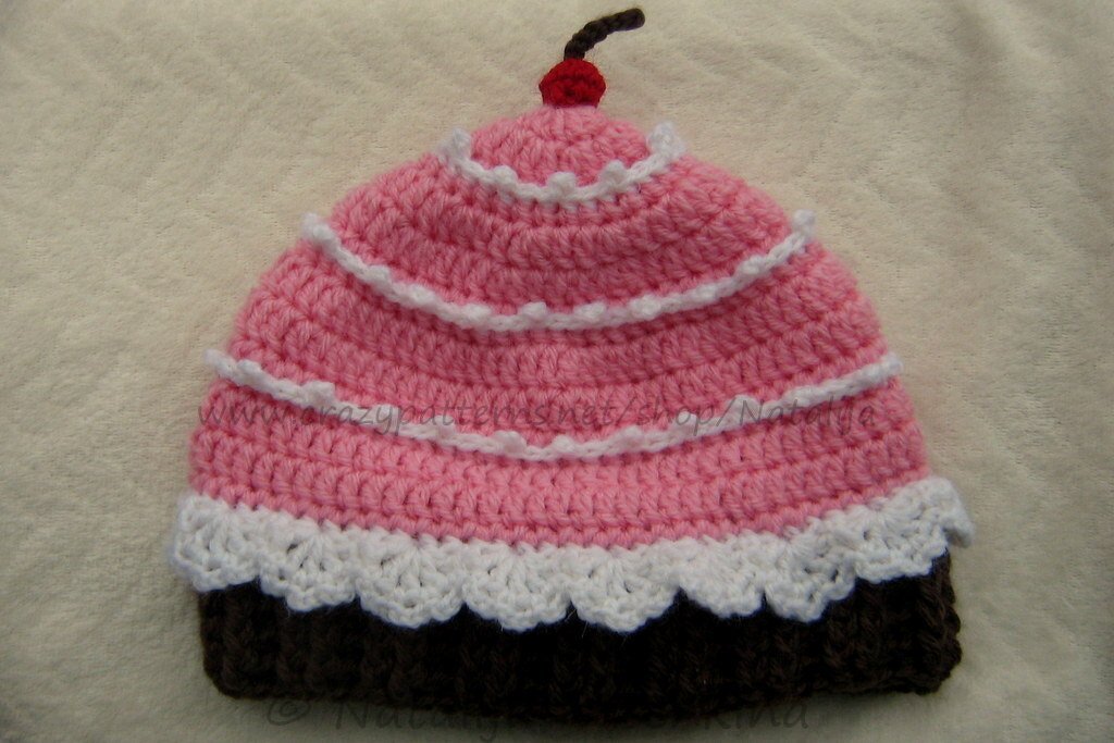 Gehäkelte Babymütze im Cupcake‑Stil mit braunem Bund, rosa Oberteil, weißen Rüschen und roter Kirsche