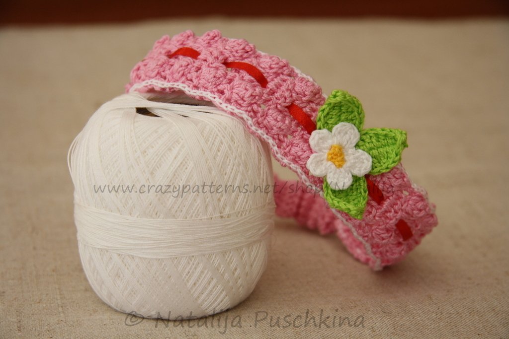 Rosa gehäkeltes Baby-Stirnband mit weißer Häkelblume, grünen Blättern und rotem Band auf weißem Garnknäuel