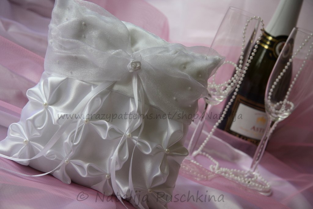 Weißes Ringkissen mit Faltornamenten, Perlenmitten und Schleifenbändern neben Gläsern