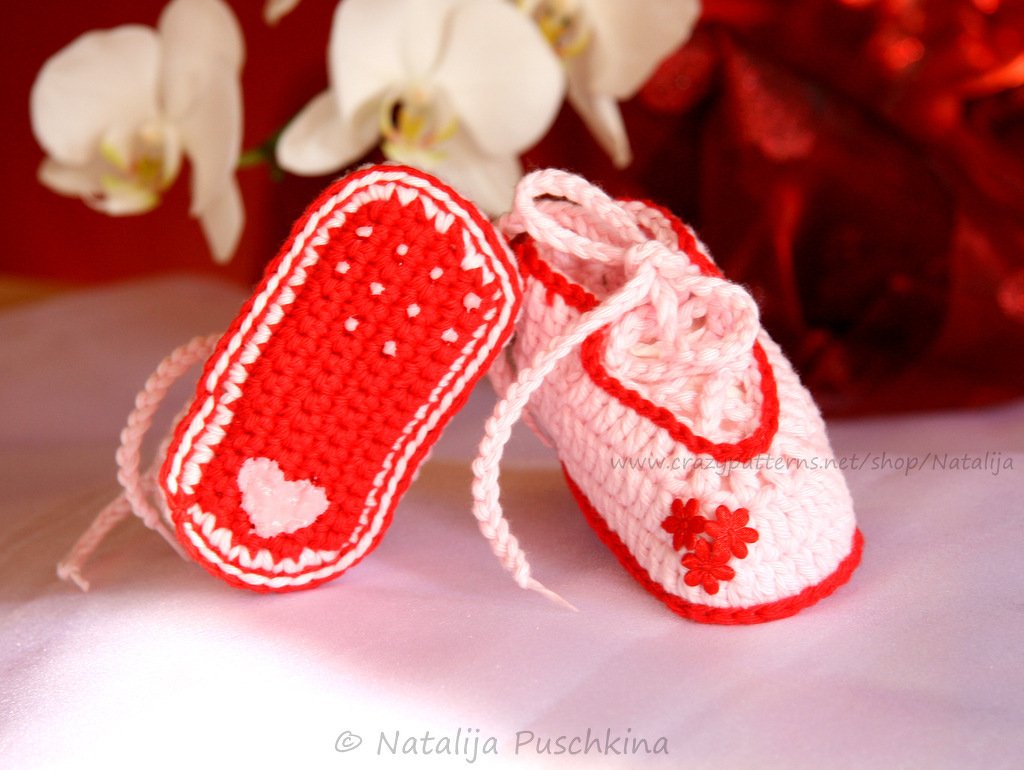 Nahaufnahme zweier gehäkelter Babyschuhe in Rosa und Rot mit Herz- und Blumenapplikationen.