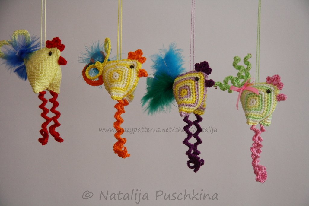 Four hanging crocheted chick ornaments in yellow, orange, purple and green with curly spiral legs and feather tails