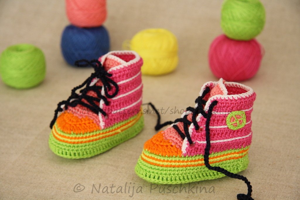 Pair of crocheted baby high-top shoes in pink, orange, green and white with black laces, with yarn balls behind them.